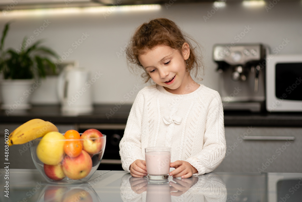 A cute little girl in a white sweater drinks yogurt. She's in the kitchen at home. Nearby, fresh fruit is on the table. The concept of healthy eating. Childhood. Taking care of the child's health. A cute little girl in a white sweater drinks yogurt. She's in the kitchen at home. Nearby, fresh fruit is on the table. The concept of healthy eating. Childhood. Taking care of the child's health.