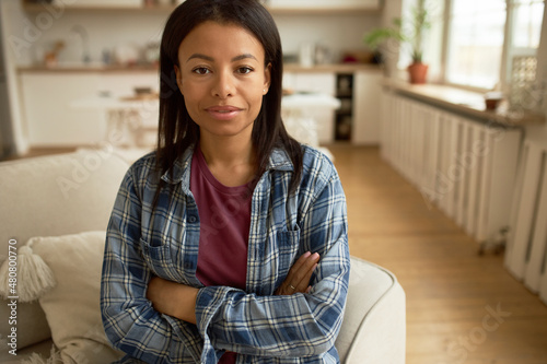 Attractive shy brunette female with brown skin sitting in closed positing with arms crossed, looking at camera, smiling, dressed casually, against home interior background. Body language