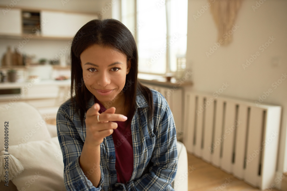 Φωτογραφία Pretty attractive dark-skinned woman of 20s sitting on couch at home, smiling pointing at you with index finger, dressed casually, having natural make-up on lovely face