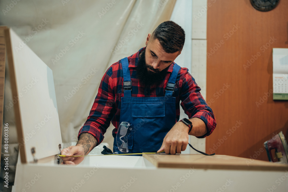Young male carpenter working in workshop and measuring with meter