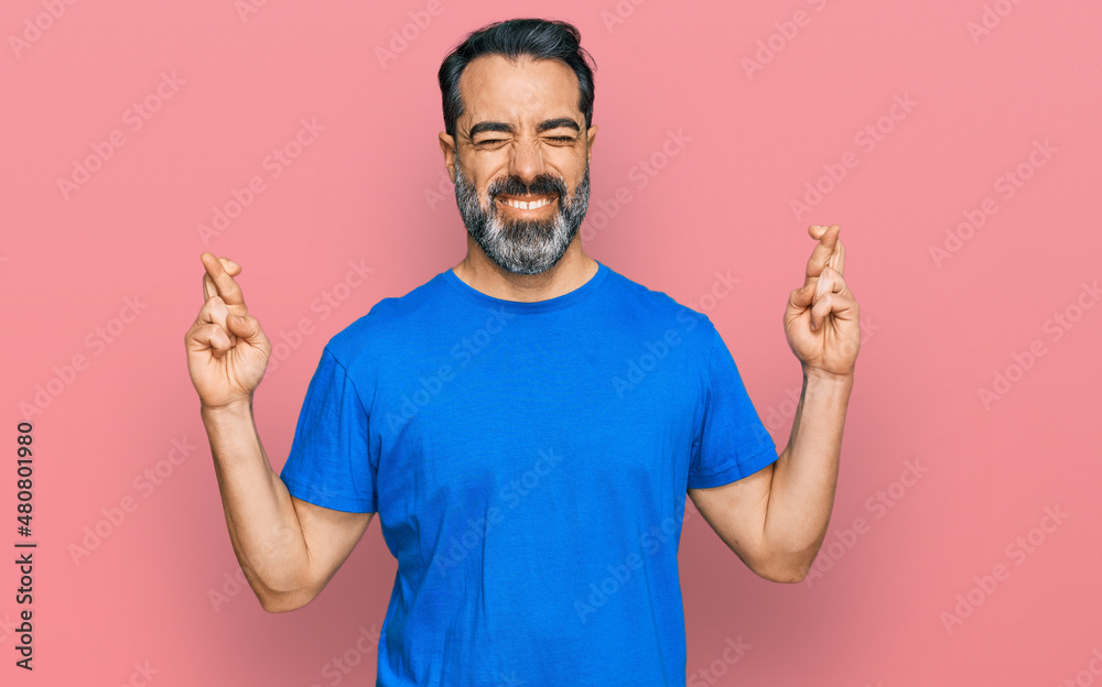 Middle aged man with beard wearing casual blue t shirt gesturing finger crossed smiling with hope and eyes closed. luck and superstitious concept.
