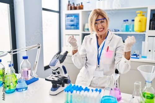 Middle age blonde woman working at scientist laboratory angry and mad raising fists frustrated and furious while shouting with anger. rage and aggressive concept.