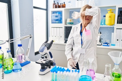 Middle age blonde woman working at scientist laboratory covering eyes with arm  looking serious and sad. sightless  hiding and rejection concept