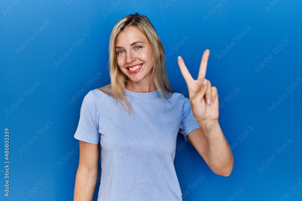 Fototapeta premium Beautiful blonde woman wearing casual t shirt over blue background smiling looking to the camera showing fingers doing victory sign. number two.
