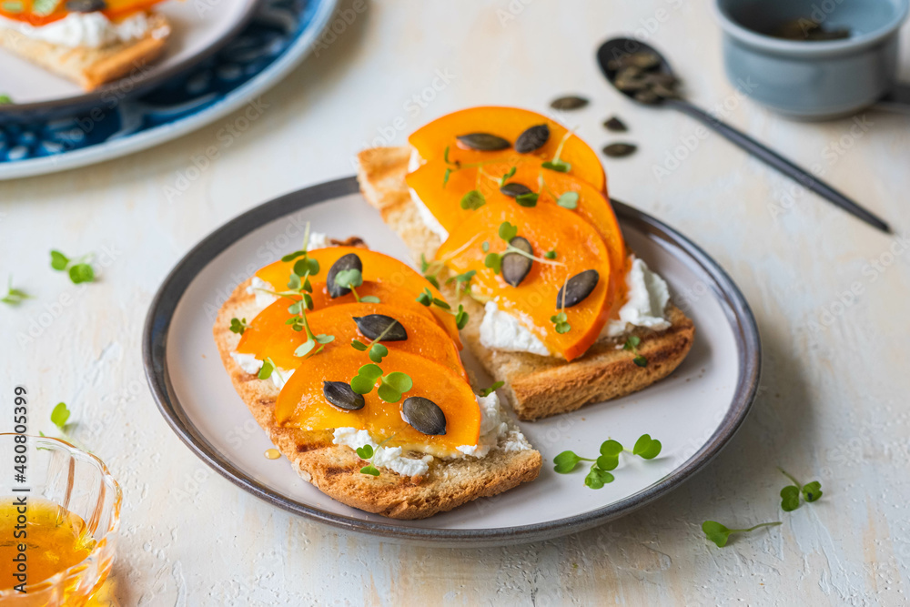 Open sandwiches with curd cheese, persimmon slices and honey on a ceramic plate on a light concrete background. Italian food. Open sandwiches with curd cheese, persimmon slices and honey on a ceramic plate on a light concrete background. Italian food.
