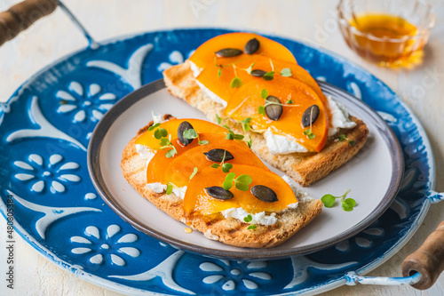 Open sandwiches with curd cheese, persimmon slices and honey on a ceramic plate on a light concrete background. Italian food.