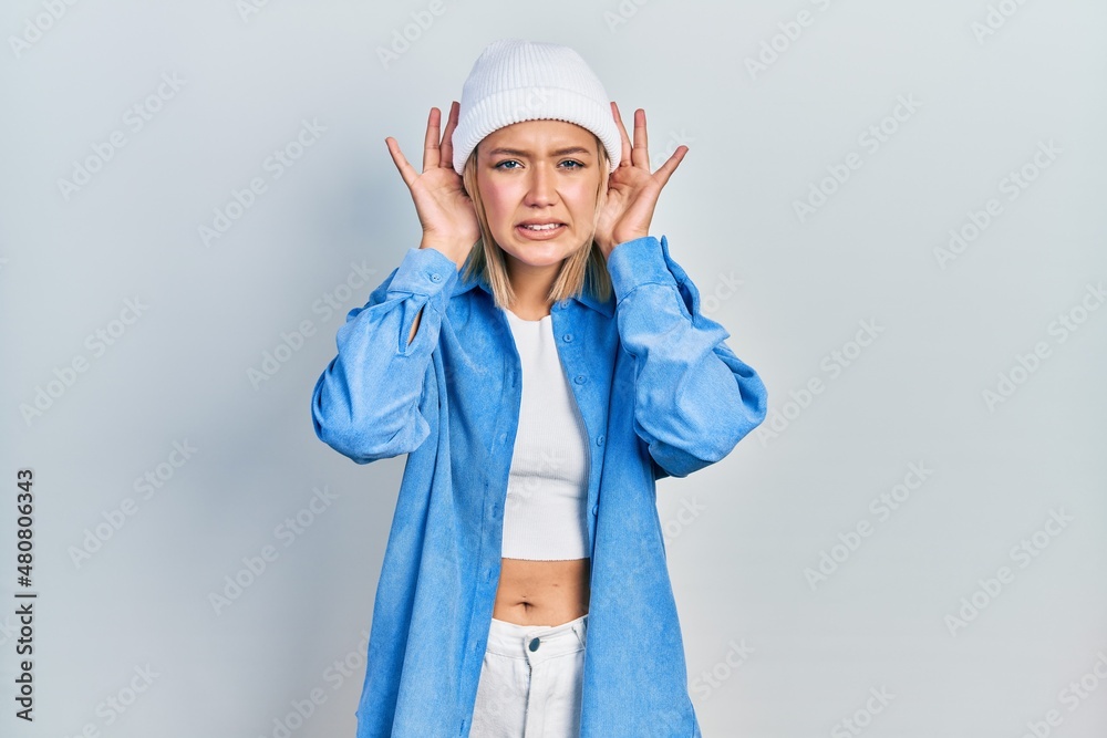 Beautiful blonde woman wearing wool hat trying to hear both hands on ear gesture, curious for gossip. hearing problem, deaf Beautiful blonde woman wearing wool hat trying to hear both hands on ear gesture, curious for gossip. hearing problem, deaf