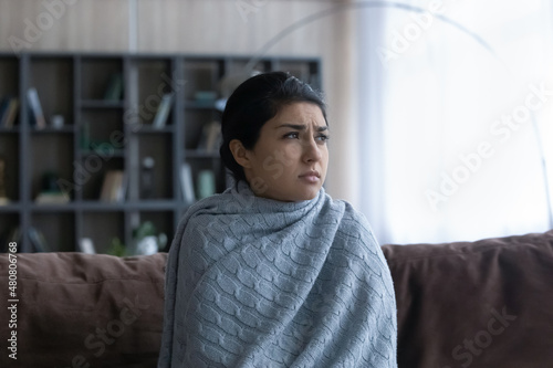 Stressed young Indian woman covered in warm plaid sitting on sofa at home, suffering from low temperature without heating system in living room or shivering having first flu symptoms.