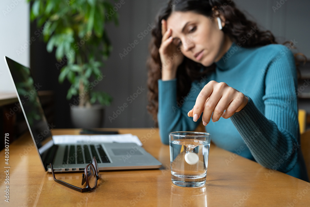Unhealthy woman suffer from headache or migraine at workplace dissolving aspirin or painkiller pill in glass of water. Sick female taking antipyretics medicine sitting alone in office, selective focus Unhealthy woman suffer from headache or migraine at workplace dissolving aspirin or painkiller pill in glass of water. Sick female taking antipyretics medicine sitting alone in office, selective focus