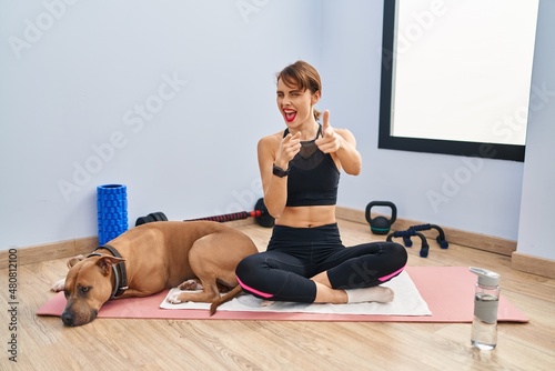 Young beautiful woman sitting on yoga mat pointing fingers to camera with happy and funny face. good energy and vibes.