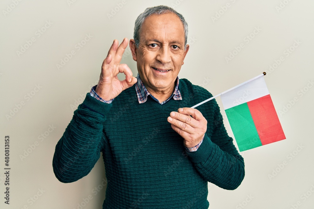 Handsome senior man with grey hair holding madagascar flag doing ok sign with fingers, smiling friendly gesturing excellent symbol Handsome senior man with grey hair holding madagascar flag doing ok sign with fingers, smiling friendly gesturing excellent symbol
