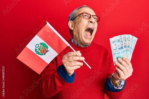 Handsome senior man with grey hair holding peru flag and peruvian sol banknotes angry and mad screaming frustrated and furious, shouting with anger looking up.