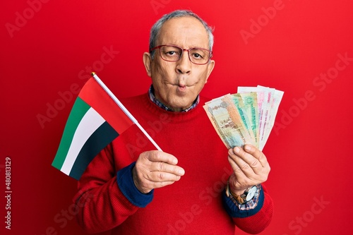 Handsome senior man with grey hair holding united arab emirates dirham banknotes making fish face with mouth and squinting eyes, crazy and comical.