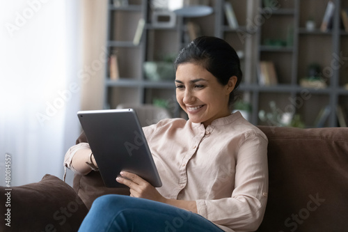 Happy young Indian woman using digital computer tablet, playing games, shopping in internet store, communicating in social networks or spending time online watching funny photo video content.