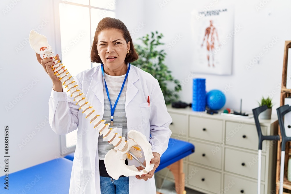 Middle age hispanic physiotherapy woman holding anatomical model of spinal column clueless and confused expression. doubt concept. Middle age hispanic physiotherapy woman holding anatomical model of spinal column clueless and confused expression. doubt concept.