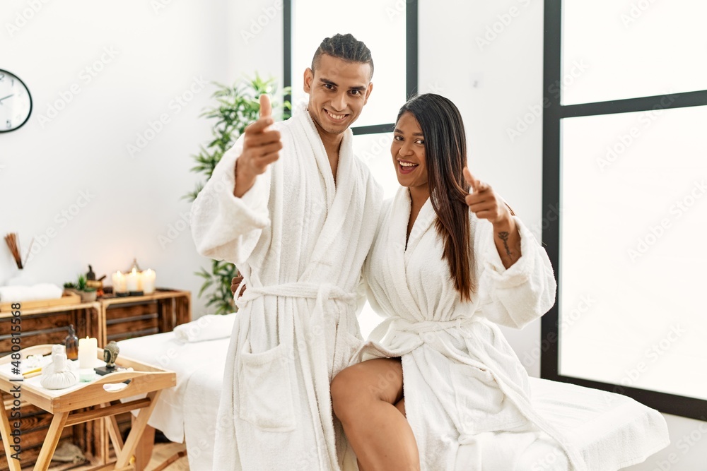 Young latin couple wearing towel standing at beauty center pointing fingers to camera with happy and funny face. good energy and vibes. Young latin couple wearing towel standing at beauty center pointing fingers to camera with happy and funny face. good energy and vibes.