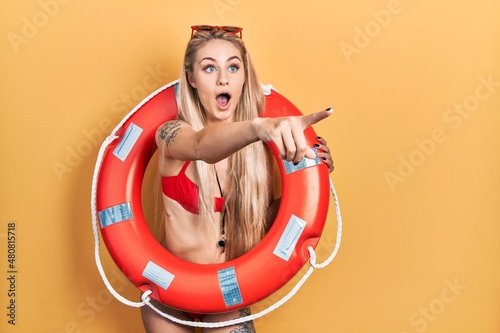 Young caucasian woman wearing bikini and holding lifeguard float pointing with finger surprised ahead, open mouth amazed expression, something on the front