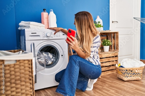 Tela Young blonde woman using smartphone and washing clothes at laundry room