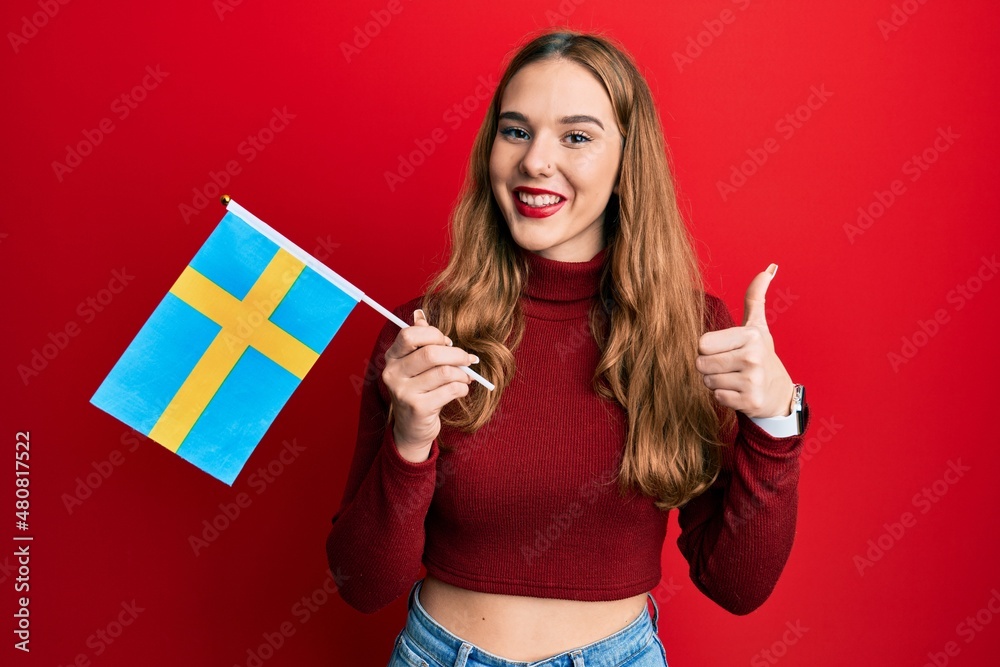 Young blonde woman holding sweden flag smiling happy and positive, thumb up doing excellent and approval sign Young blonde woman holding sweden flag smiling happy and positive, thumb up doing excellent and approval sign