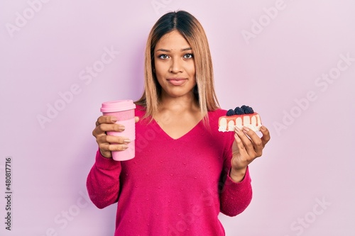 Tableau sur toile Beautiful hispanic woman holding takeaway cup of coffee and cheesecake relaxed with serious expression on face
