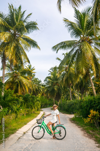 Girl is cycling on her old vintage bicycle through the jungle of Anse Source d’Argent, la Digue, Seychelles. She is surrounded by palm trees and flowers. Enjoying her beautiful holiday.