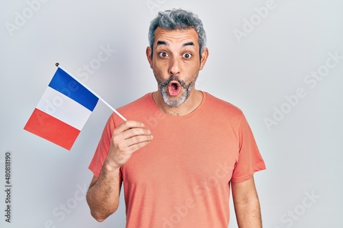 Handsome middle age man with grey hair holding france flag scared and amazed with open mouth for surprise, disbelief face