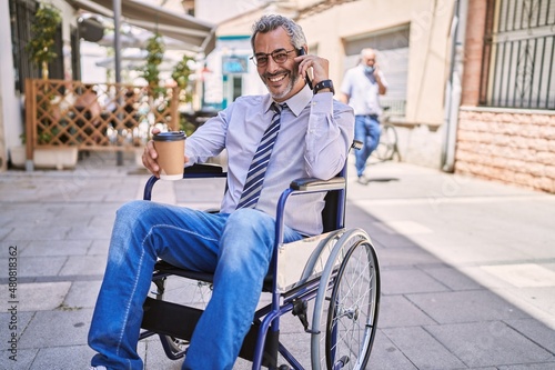 Middle age hispanic man sitting on wheelchair talking on the smartphone at street