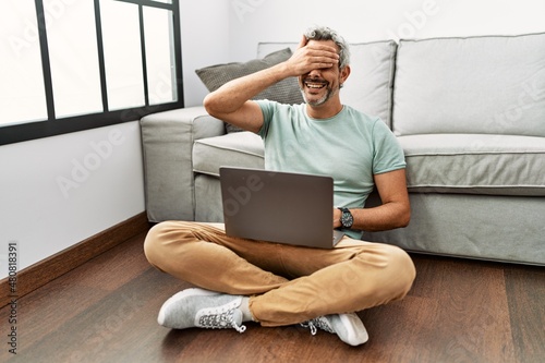 Middle age hispanic man using laptop sitting on the floor at the living room smiling and laughing with hand on face covering eyes for surprise. blind concept.