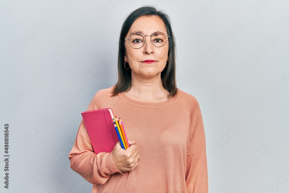 Middle age hispanic woman holding book and color pencils thinking attitude and sober expression looking self confident Middle age hispanic woman holding book and color pencils thinking attitude and sober expression looking self confident