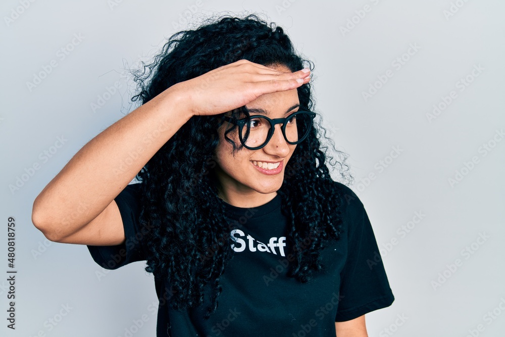 Young hispanic woman with curly hair wearing staff t shirt very happy and smiling looking far away with hand over head. searching concept. Young hispanic woman with curly hair wearing staff t shirt very happy and smiling looking far away with hand over head. searching concept.