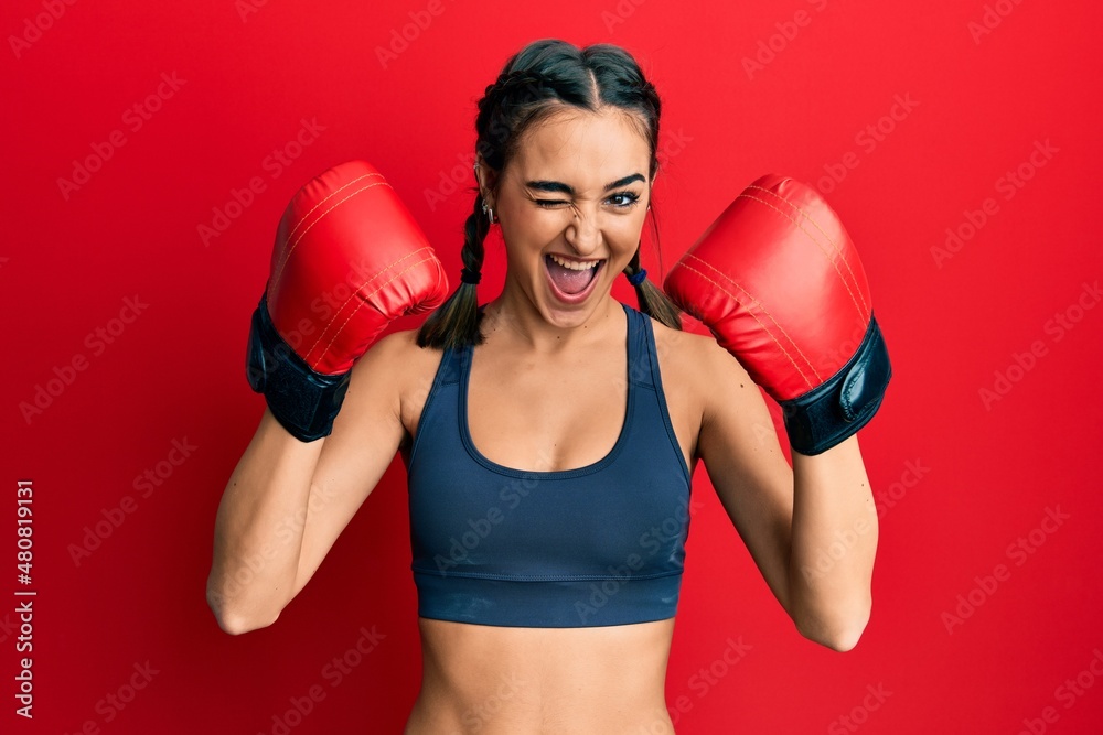 Young brunette girl using boxing gloves wearing braids winking looking at the camera with sexy expression, cheerful and happy face.