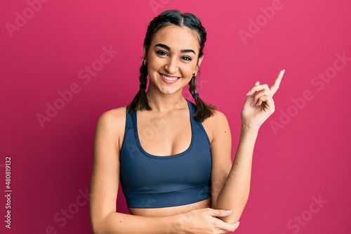 Young brunette girl wearing sportswear and braids with a big smile on face  pointing with hand and finger to the side looking at the camera.