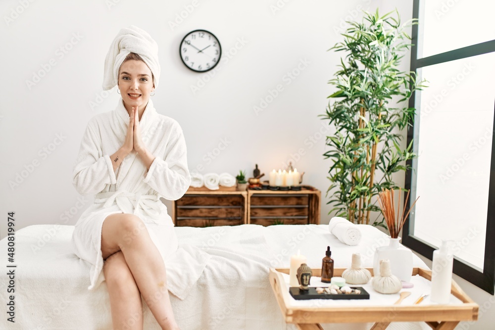 Young blonde woman wearing bathrobe at wellbeing spa praying with hands together asking for forgiveness smiling confident. Young blonde woman wearing bathrobe at wellbeing spa praying with hands together asking for forgiveness smiling confident.