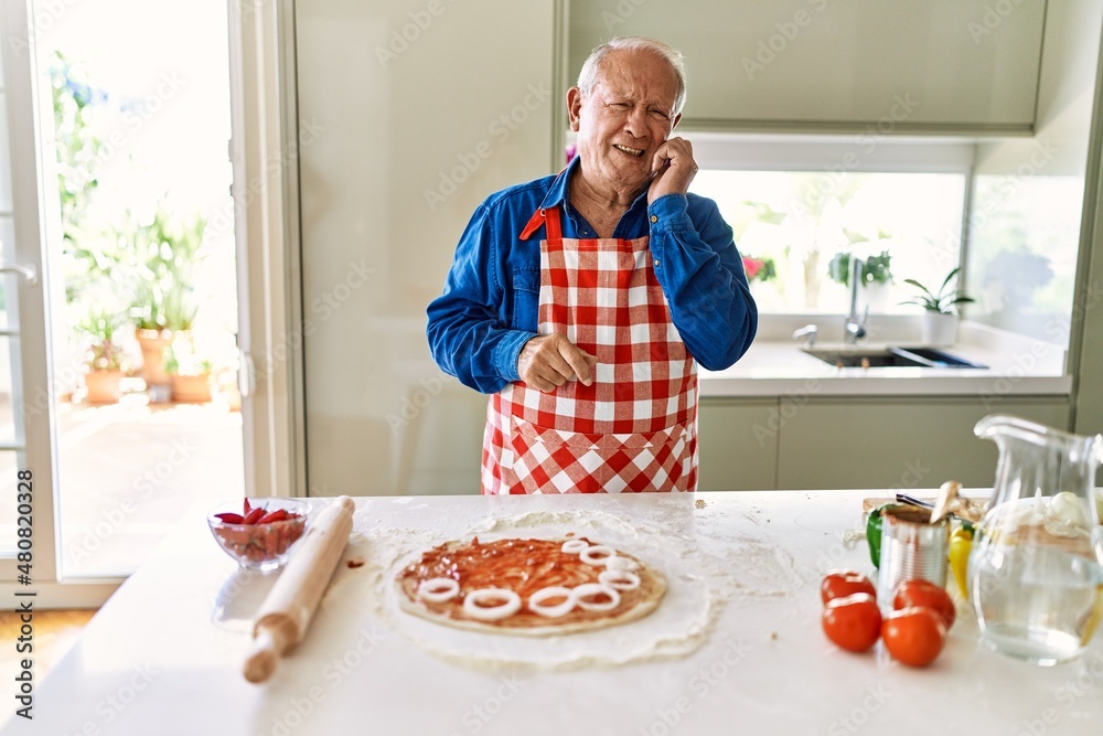 Senior man with grey hair cooking pizza at home kitchen looking stressed and nervous with hands on mouth biting nails. anxiety problem. Senior man with grey hair cooking pizza at home kitchen looking stressed and nervous with hands on mouth biting nails. anxiety problem.