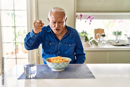 Senior man with grey hair eating pasta spaghetti at home angry and mad raising fist frustrated and furious while shouting with anger. rage and aggressive concept.