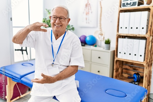 Senior physiotherapy man working at pain recovery clinic gesturing with hands showing big and large size sign  measure symbol. smiling looking at the camera. measuring concept.