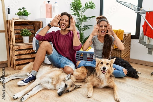 Photography Young hispanic couple doing laundry with dogs smiling cheerful playing peek a boo with hands showing face
