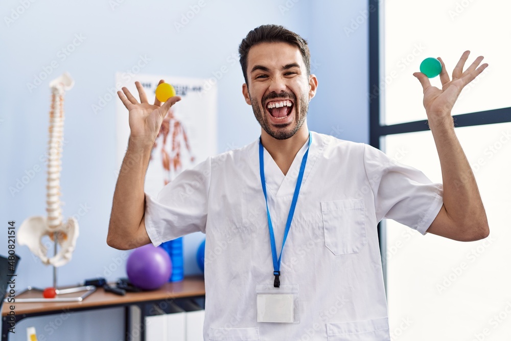 Young hispanic physiotherapist man holding strength balls to train hand muscles celebrating crazy and amazed for success with open eyes screaming excited. Young hispanic physiotherapist man holding strength balls to train hand muscles celebrating crazy and amazed for success with open eyes screaming excited.