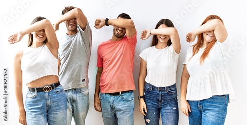 Group of young friends standing together over isolated background covering eyes with arm smiling cheerful and funny. blind concept.