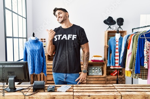 Young hispanic man working at retail boutique smiling with happy face looking and pointing to the side with thumb up.