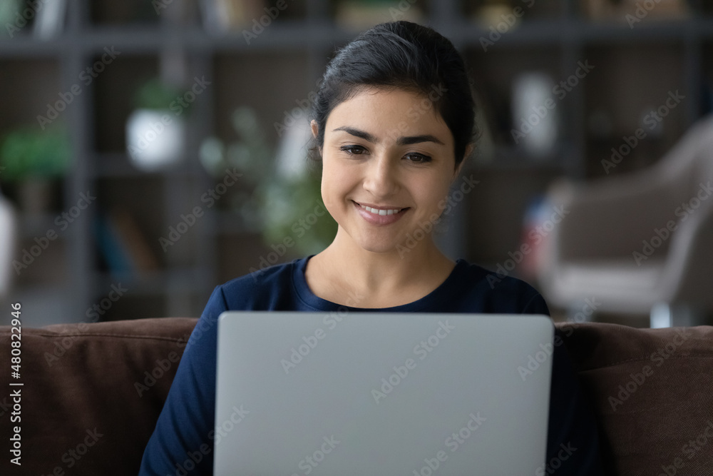 Happy attractive millennial Indian ethnicity woman working on computer, typing message enjoying communicating in social network, choosing goods shopping in internet store sitting on sofa at home.