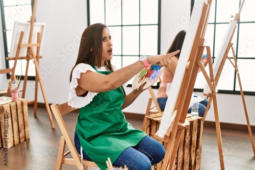 Young hispanic artist women painting on canvas at art studio pointing with finger surprised ahead, open mouth amazed expression, something on the front