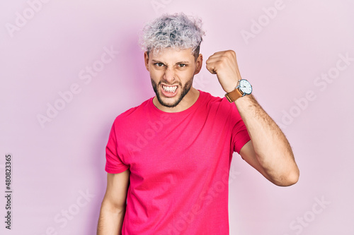Young hispanic man with modern dyed hair wearing casual pink t shirt angry and mad raising fist frustrated and furious while shouting with anger. rage and aggressive concept.