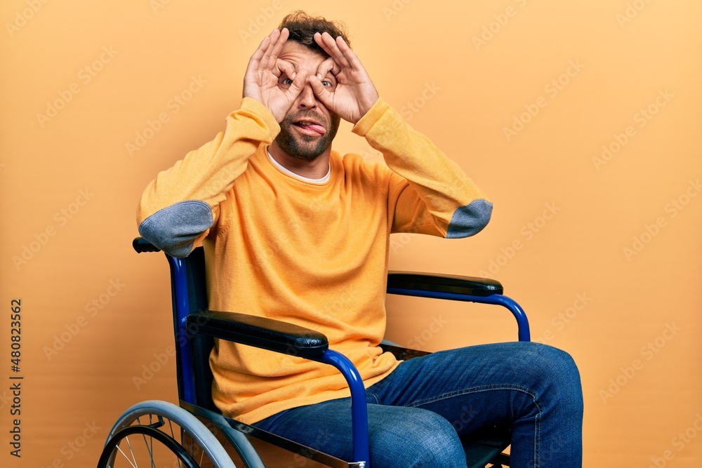 Handsome man with beard sitting on wheelchair doing ok gesture like binoculars sticking tongue out, eyes looking through fingers. crazy expression. Handsome man with beard sitting on wheelchair doing ok gesture like binoculars sticking tongue out, eyes looking through fingers. crazy expression.