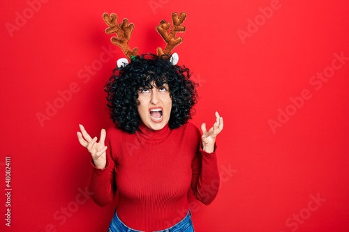 Young middle east woman wearing cute christmas reindeer horns crazy and mad shouting and yelling with aggressive expression and arms raised. frustration concept.