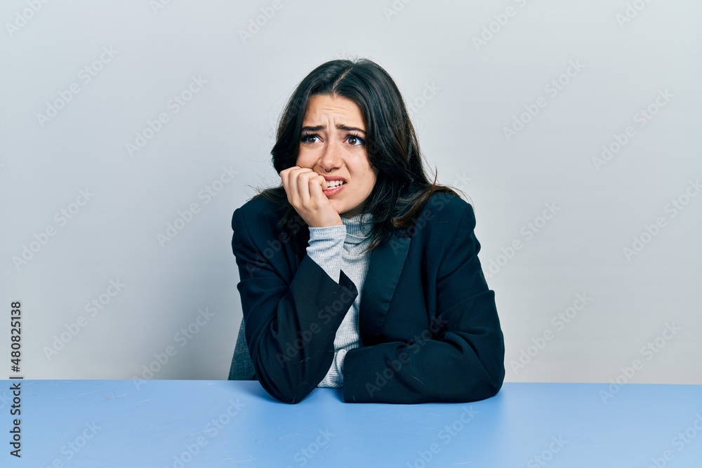 Beautiful hispanic business woman sitting on the table looking stressed and nervous with hands on mouth biting nails. anxiety problem. Beautiful hispanic business woman sitting on the table looking stressed and nervous with hands on mouth biting nails. anxiety problem.