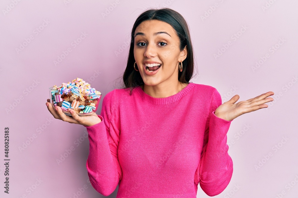 Young hispanic woman holding bowl of sugar candy celebrating achievement with happy smile and winner expression with raised hand Young hispanic woman holding bowl of sugar candy celebrating achievement with happy smile and winner expression with raised hand