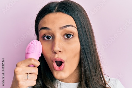 Young hispanic woman using facial exfoliating cleaner scared and amazed with open mouth for surprise  disbelief face