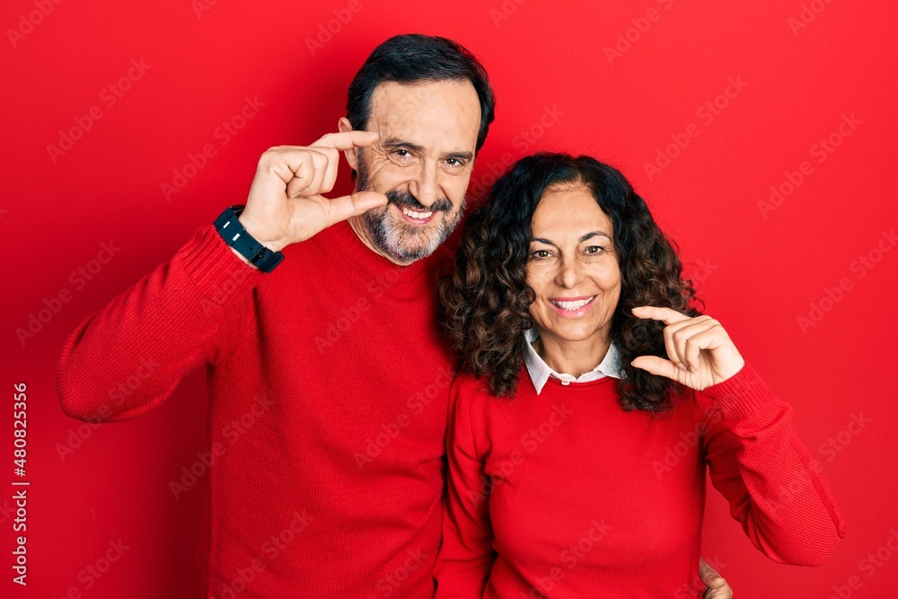 Middle age couple of hispanic woman and man hugging and standing together smiling and confident gesturing with hand doing small size sign with fingers looking and the camera. measure concept. Middle age couple of hispanic woman and man hugging and standing together smiling and confident gesturing with hand doing small size sign with fingers looking and the camera. measure concept.