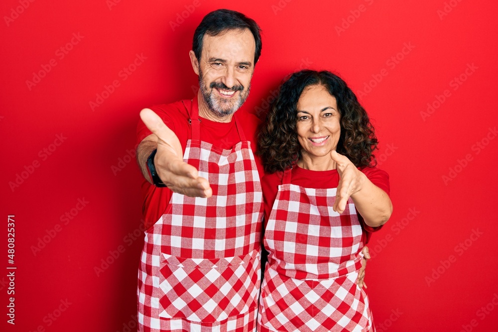Middle age couple of hispanic woman and man wearing cook apron smiling friendly offering handshake as greeting and welcoming. successful business. Middle age couple of hispanic woman and man wearing cook apron smiling friendly offering handshake as greeting and welcoming. successful business.